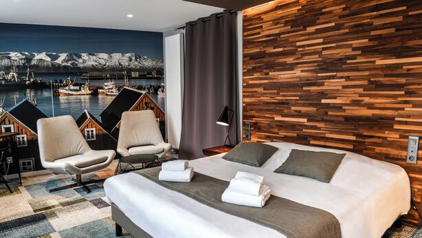 A hotel room with a large bed, two armchairs, a view of a harbor with boats, and a wooden wall. - Le K'Loys (Paimpol)