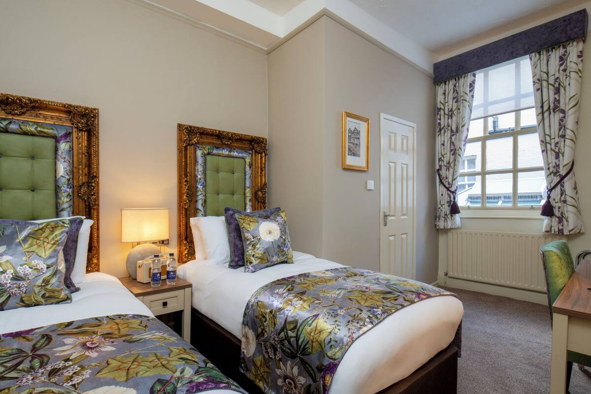 A hotel room with two beds, a nightstand, a lamp, a window with curtains, and a radiator.