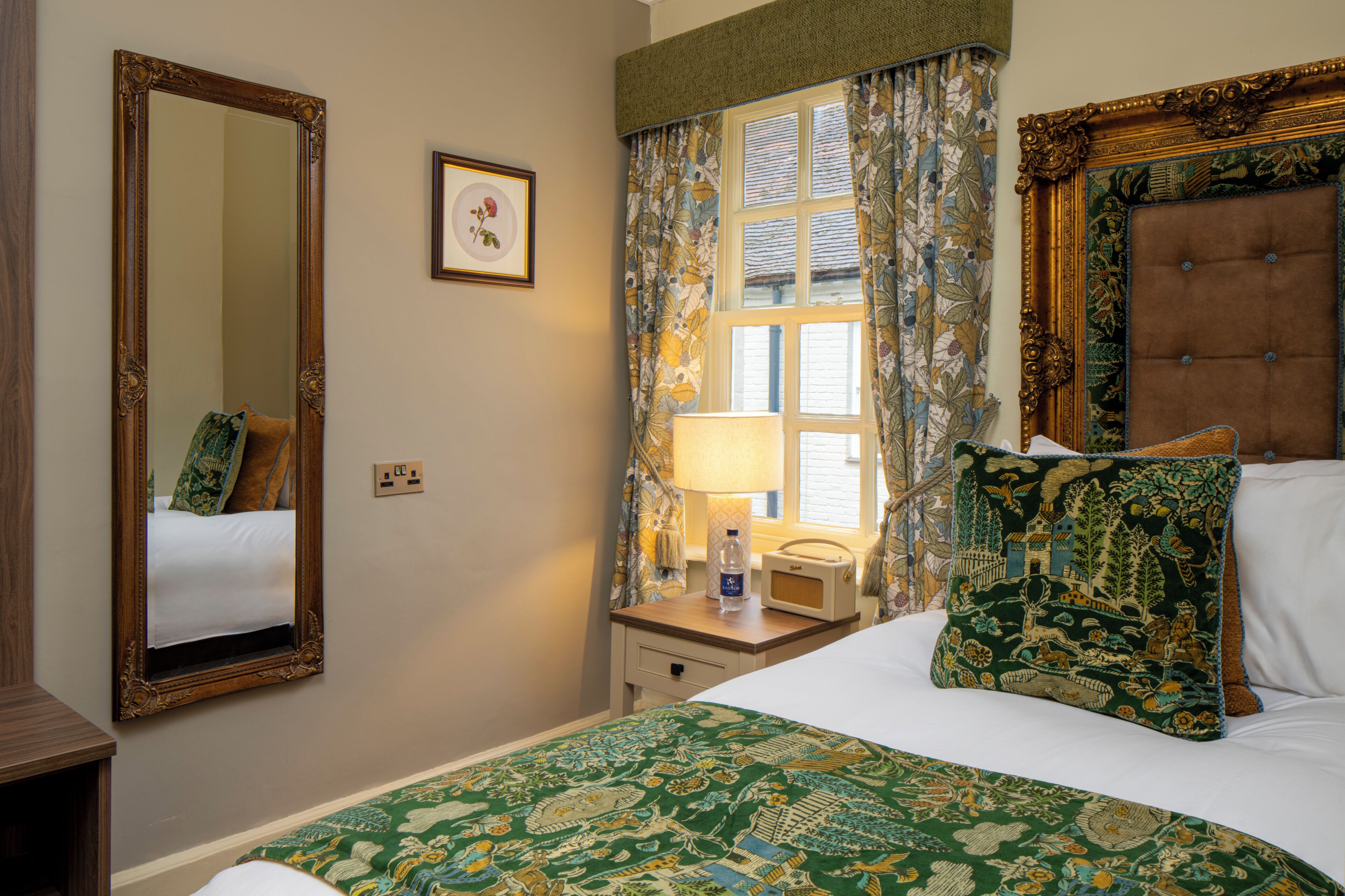 A hotel room with a large mirror, a bed with a green patterned bedspread, a window with floral curtains, and a bedside table with a lamp and a bottle.