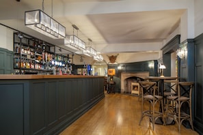 Una zona de bar con suelo de madera, barra con estantes llenos de botellas y chimenea con asientos.