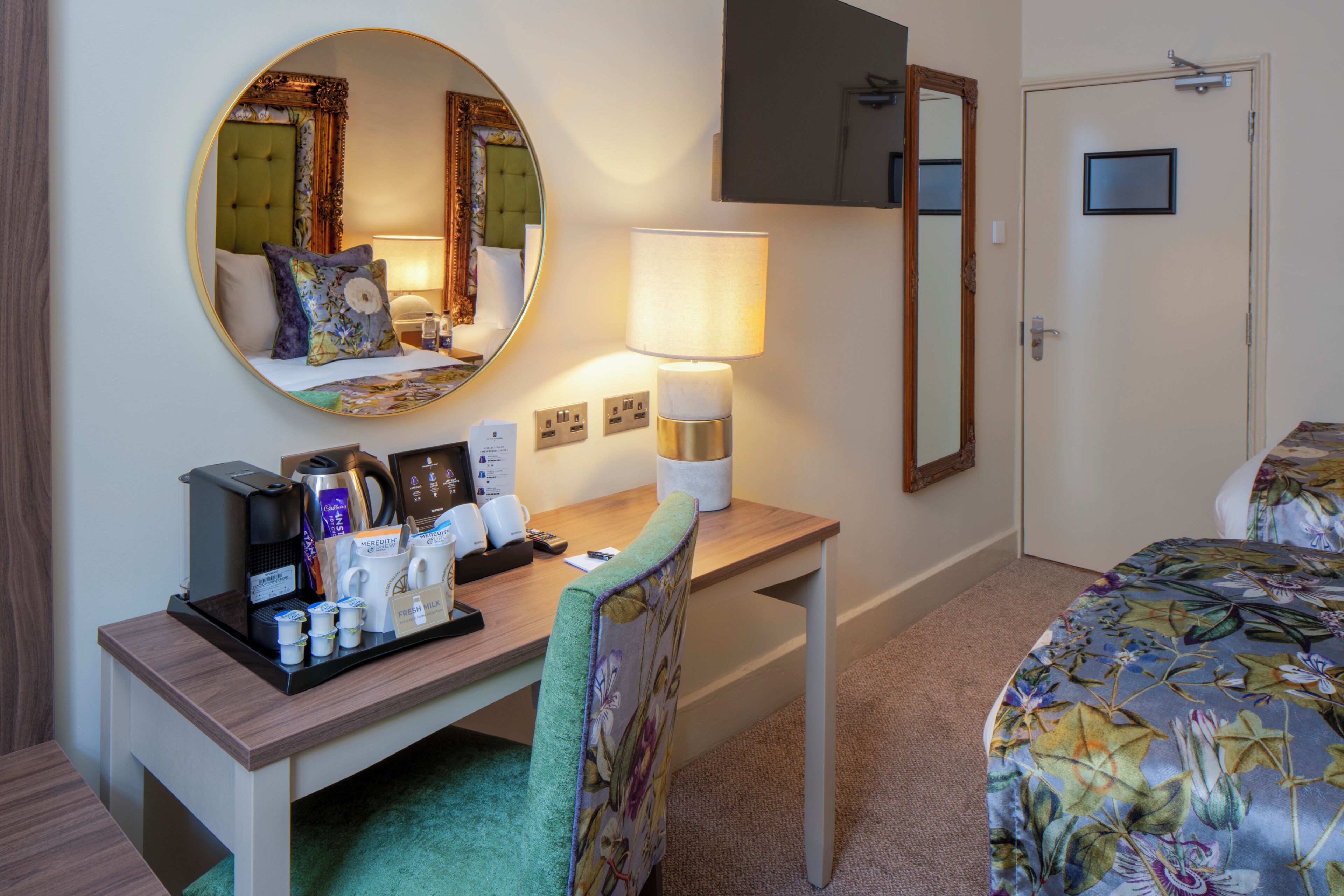 A hotel room with a bed, a desk with a lamp, a mirror, and a coffee maker.