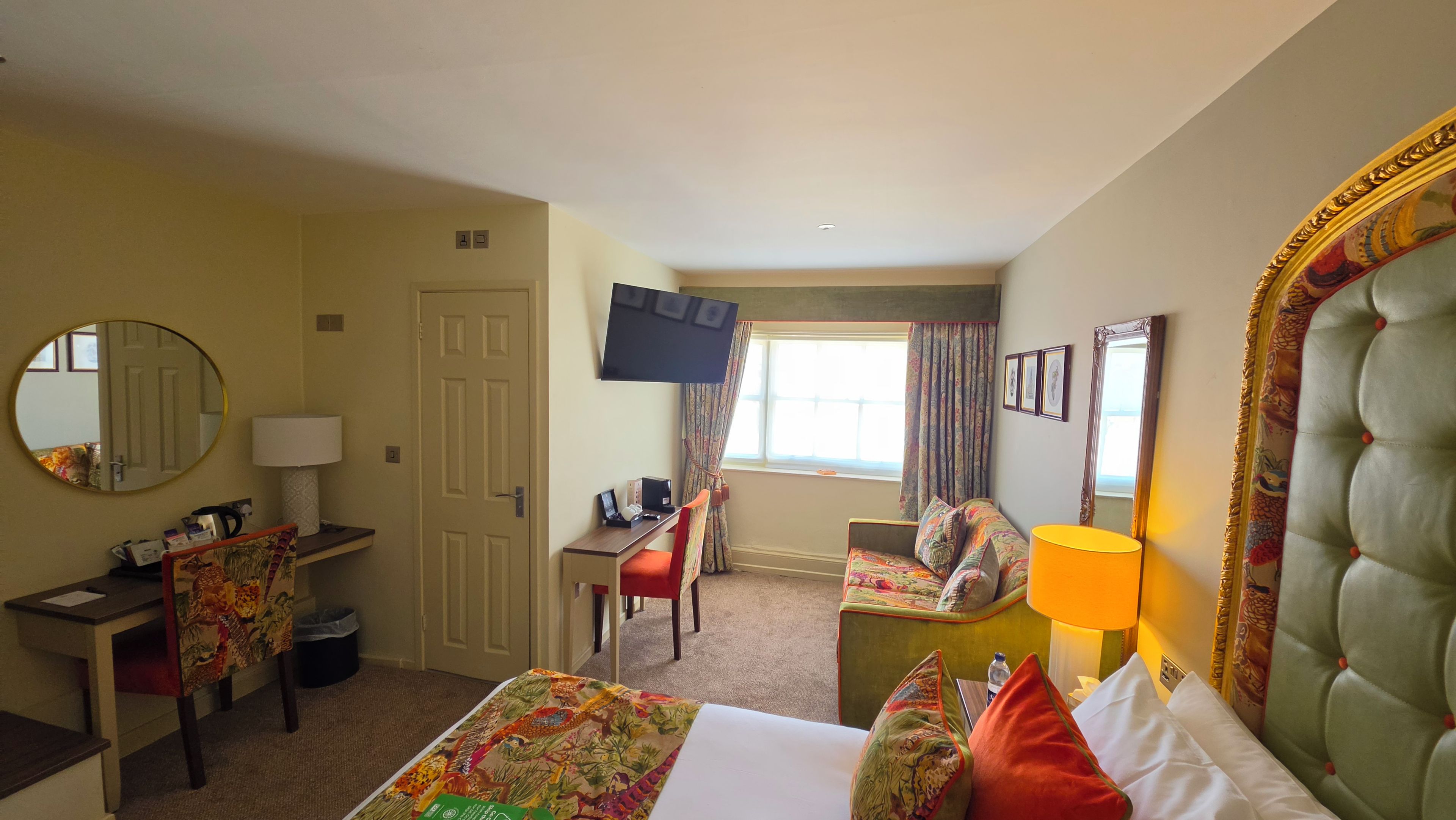 Habitación de hotel con cama, escritorio, silla, televisión, ventana con cortinas y espejo.
