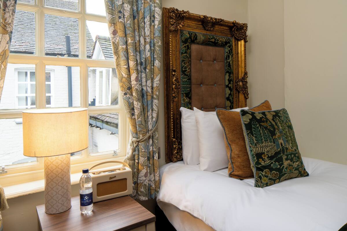 A hotel room with a bed, a bedside table with a lamp and a bottle, a window with curtains, and a decorative headboard.