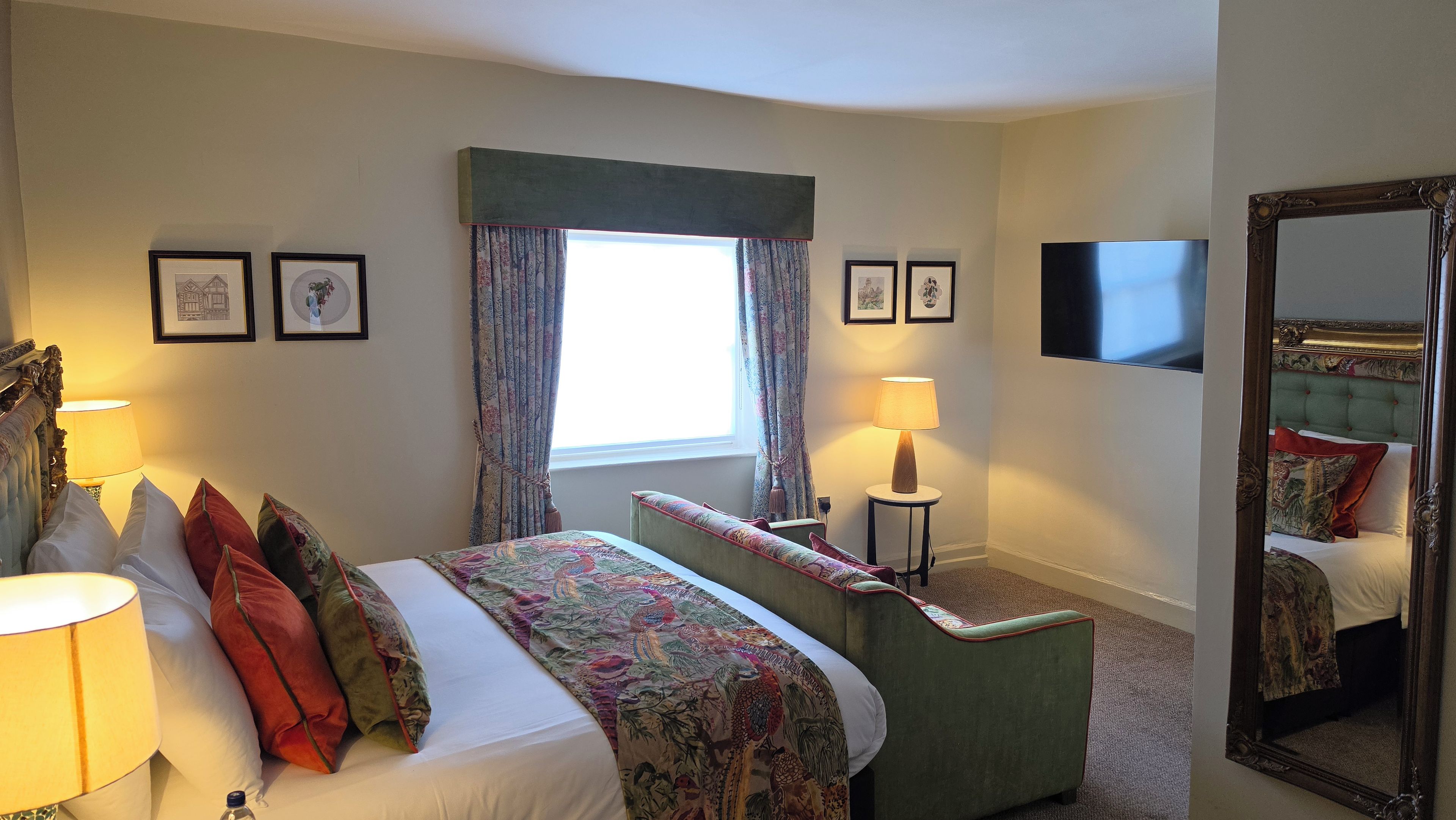 A hotel room with a bed, bedside table, lamp, window with curtains, framed pictures on the wall, and a large mirror.