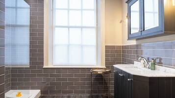 A bathroom with dark tiled walls, a window, a sink with a mirror, and a small stool.