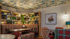A cozy interior with a wooden bench, a round table, and patterned chairs. Shelves with decorative items and a framed picture are also visible.