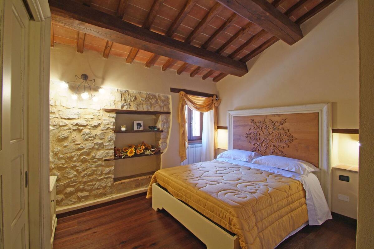 A bedroom with a stone wall, wooden beams, a bed with a headboard, and a shelf with decorative items.