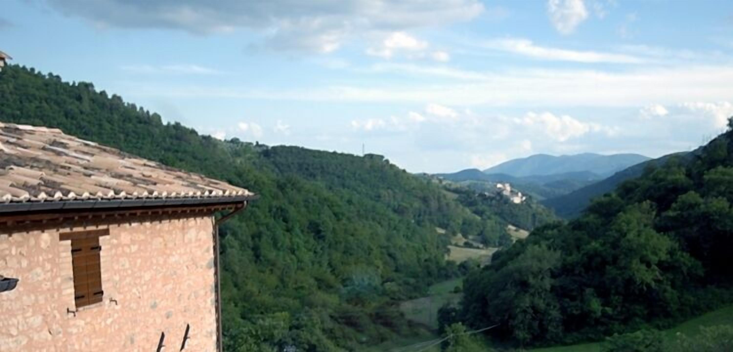 A rustic stone building with a tiled roof, surrounded by lush green hills and distant mountains.