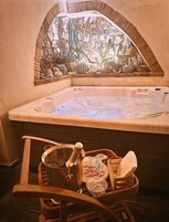 Un jacuzzi con pared de piedra y una ventana encima. Hay una silla de madera, un cubo y una toalla al lado de la bañera.