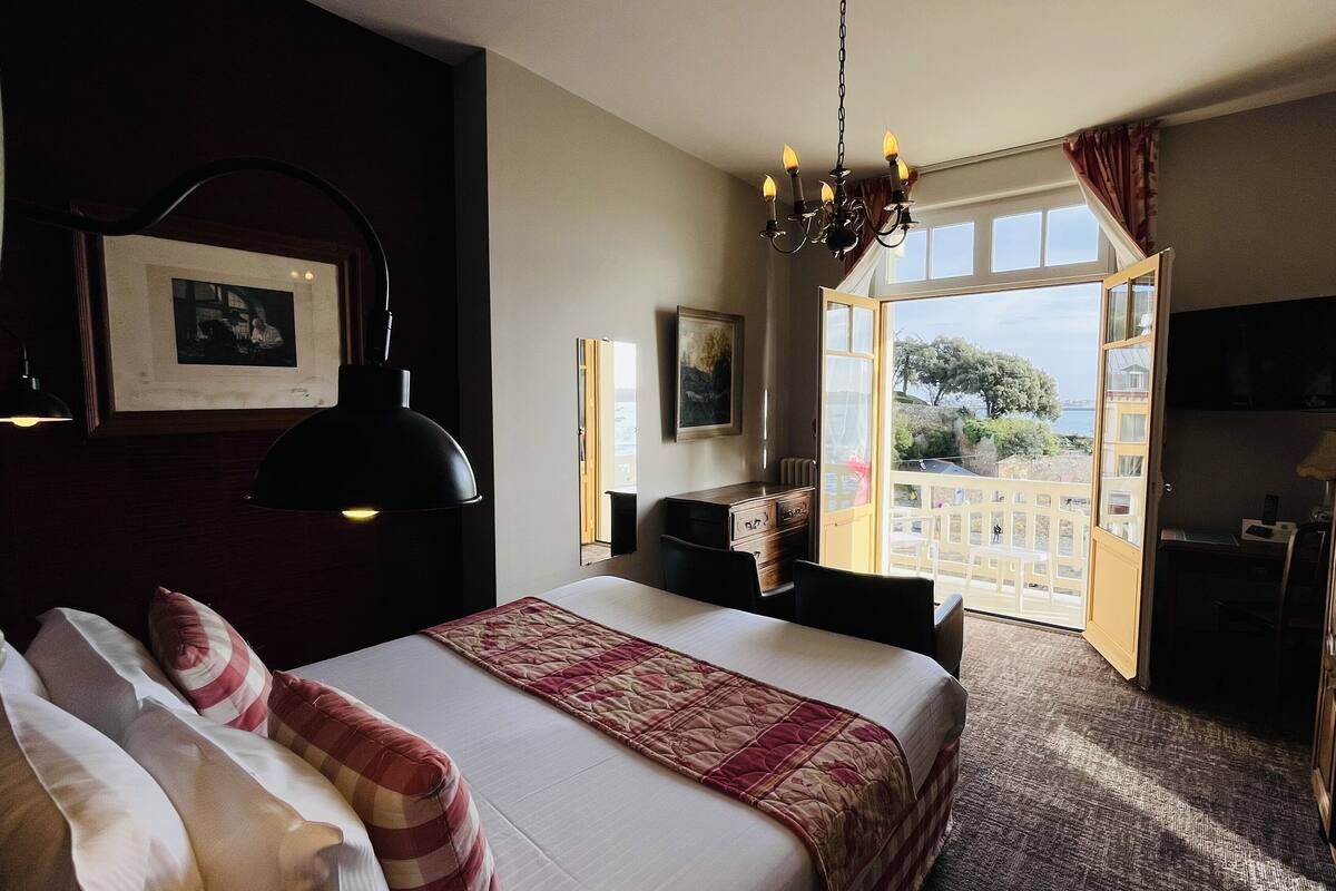 A hotel room with a bed, a desk, a chair, a wardrobe, and a large window with a view of the sea.