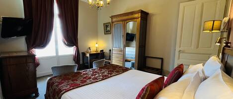 A hotel room with a large bed, a desk, a chair, a television, a wardrobe, and a chandelier.