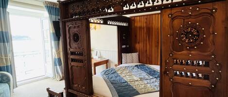 A wooden four-poster bed with intricate carvings, a bedside table with a lamp, and a view of the outdoors through a large window.