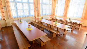 A dining area with multiple wooden tables and benches, arranged in a spacious room with large windows and curtains.