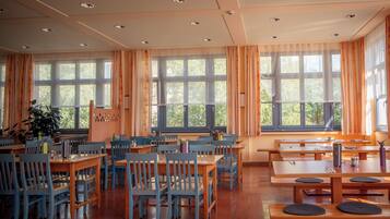 A spacious dining area with multiple wooden tables and chairs, large windows with curtains, and a ceiling with recessed lighting.