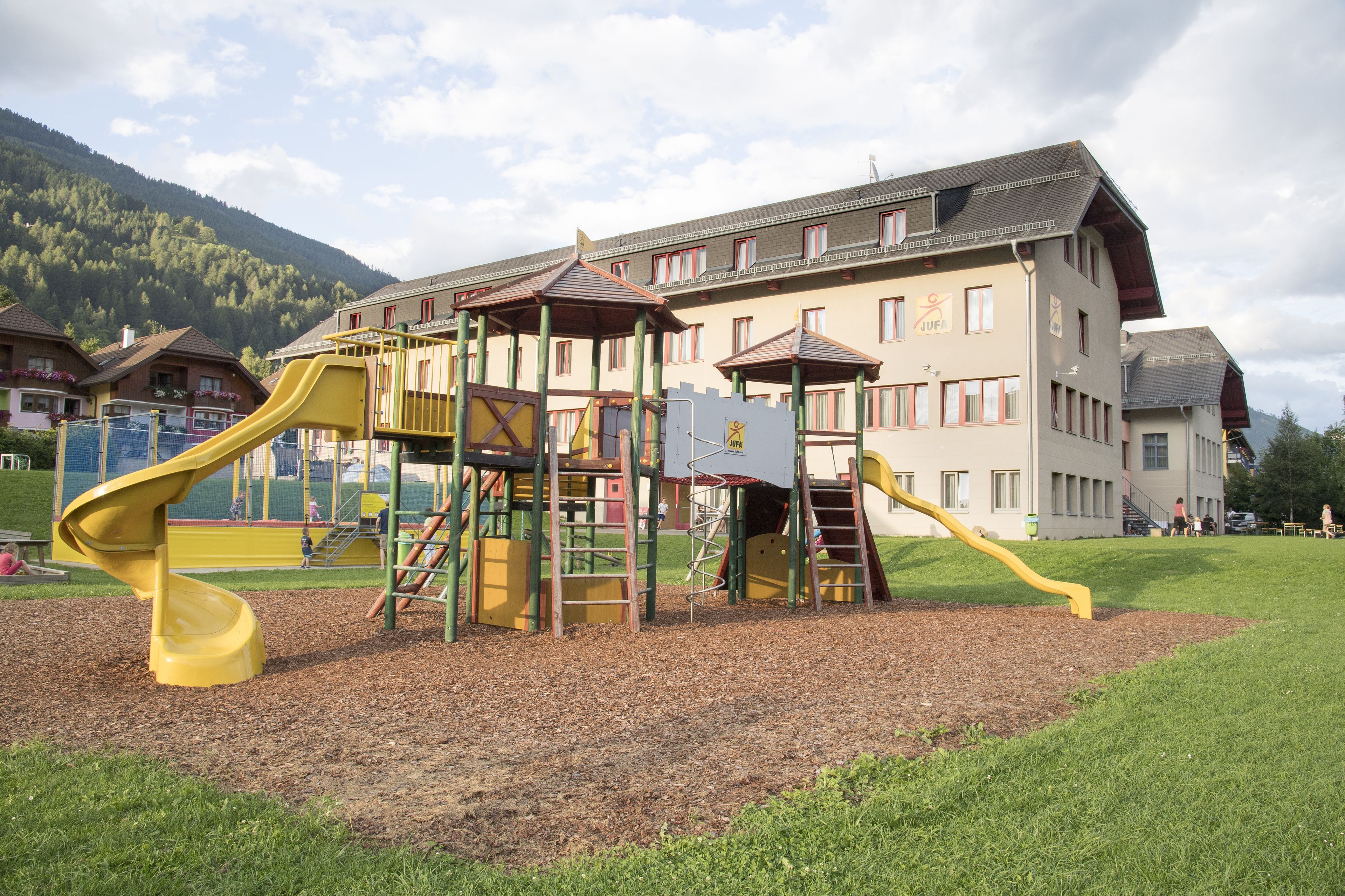 Un’area giochi con uno scivolo giallo e strutture di arrampicata, sullo sfondo di un grande edificio con molte finestre e una montagna all’orizzonte.