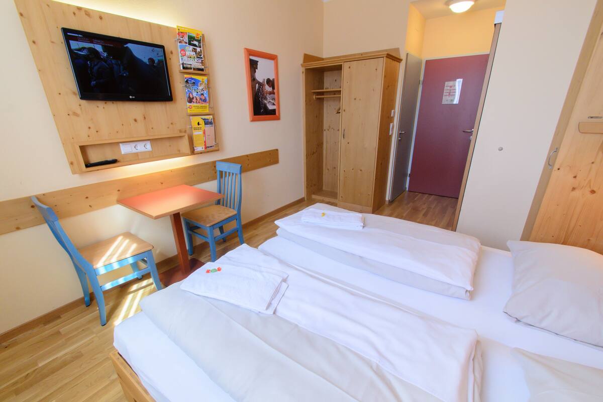 Ein Hotelzimmer mit einem an der Wand befestigten Fernseher, einem Holzschrank, einem Schreibtisch mit Stuhl und zwei Betten mit weißen Bettwäsche.