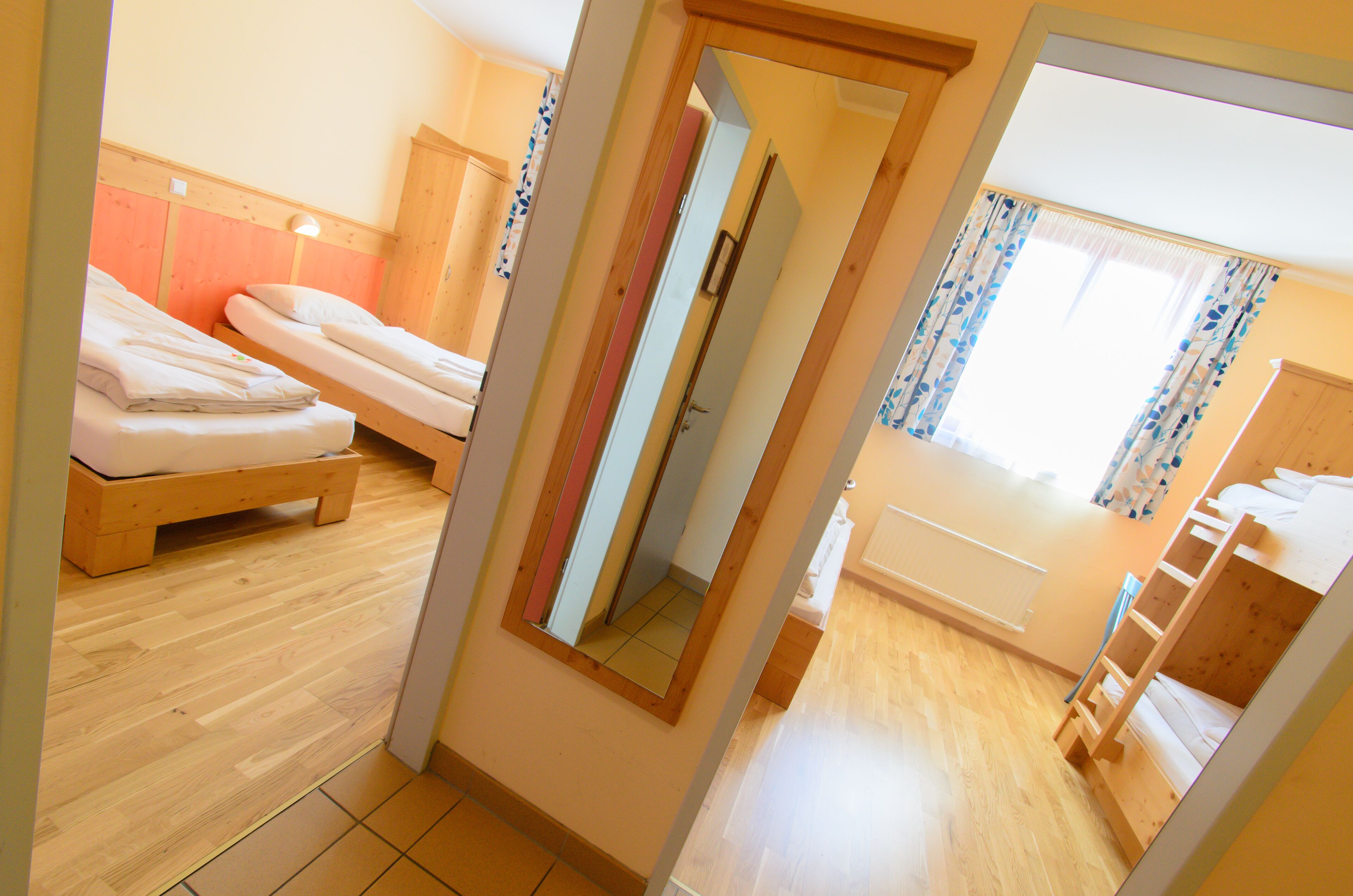 A room with two wooden bunk beds, a wooden wardrobe, a mirror, and a window with curtains.