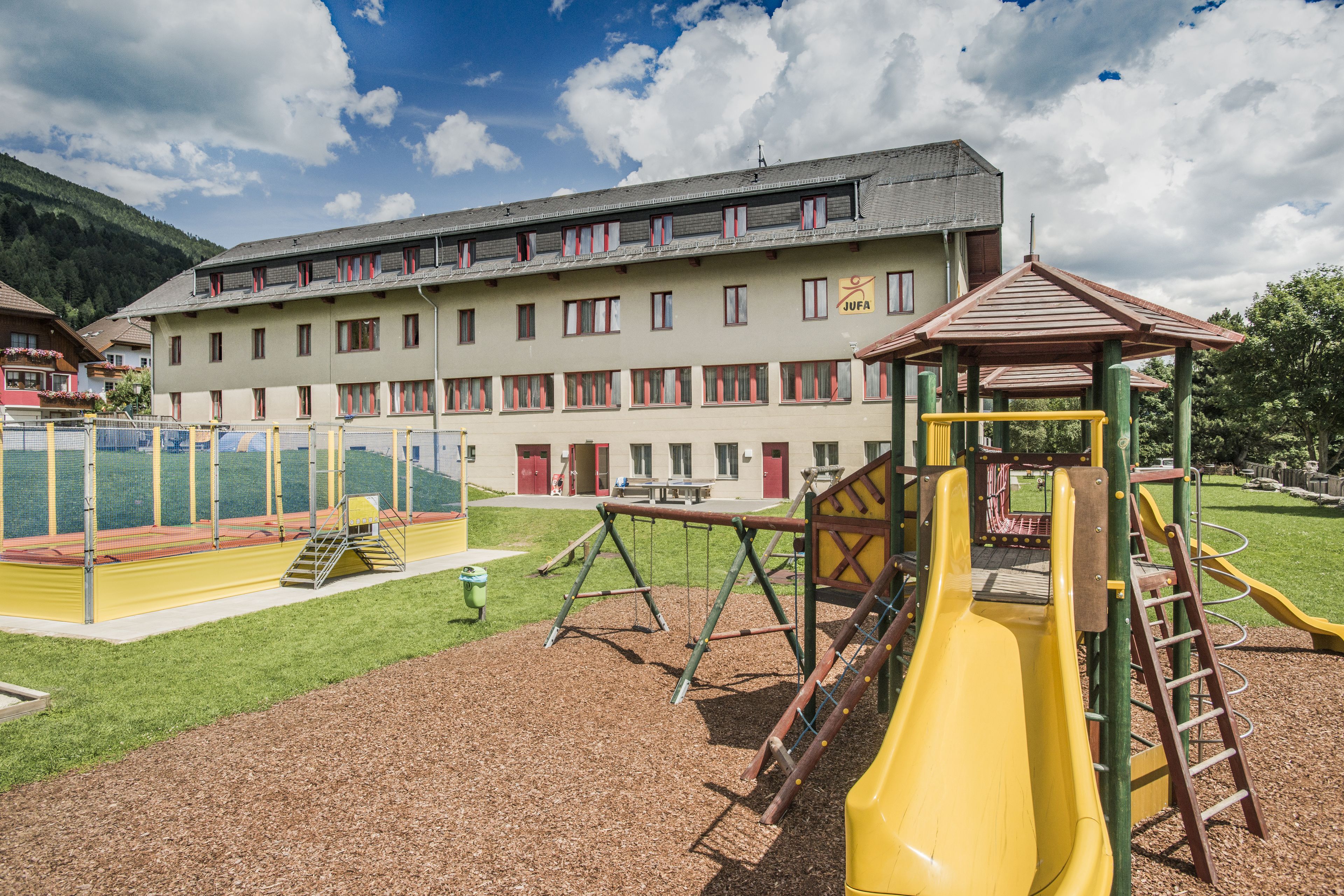 A playground with a yellow slide and swings, surrounded by a building with multiple windows and a red door.