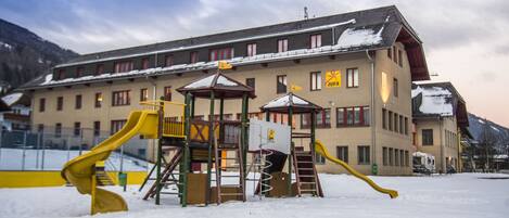 Un parque infantil con toboganes y una estructura de escalada frente a un edificio con múltiples ventanas y un tejado cubierto de nieve.