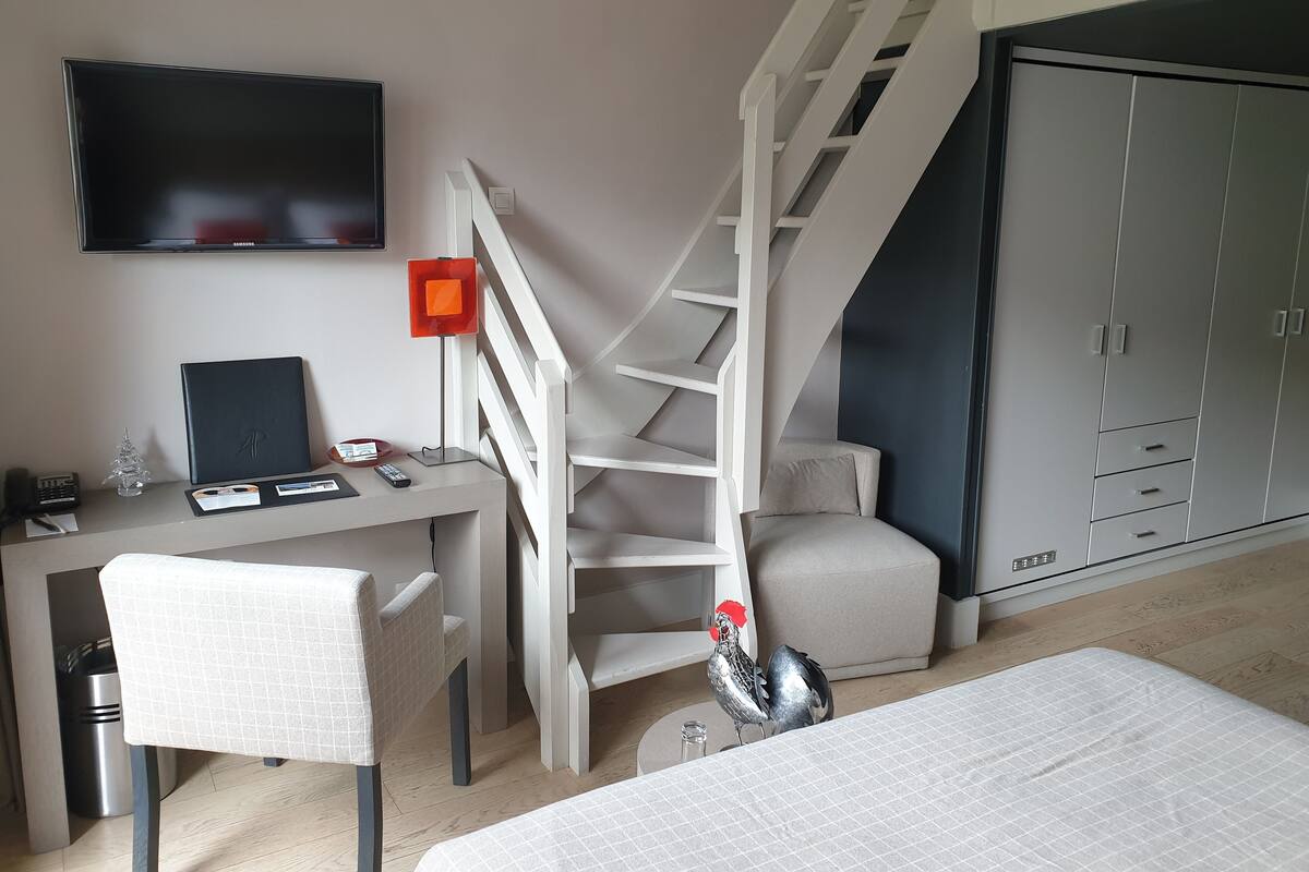 A bedroom with a bed, a desk with a computer, a chair, a TV, a staircase, and a wardrobe.