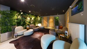 A modern lounge area with a dark ceiling, recessed lighting, and a variety of seating options including white armchairs and ottomans. There are several potted plants adding greenery to the space, and a large window with a view of the outdoors. - Hotel & Spa L'Alta Peyra (Saint-Veran)