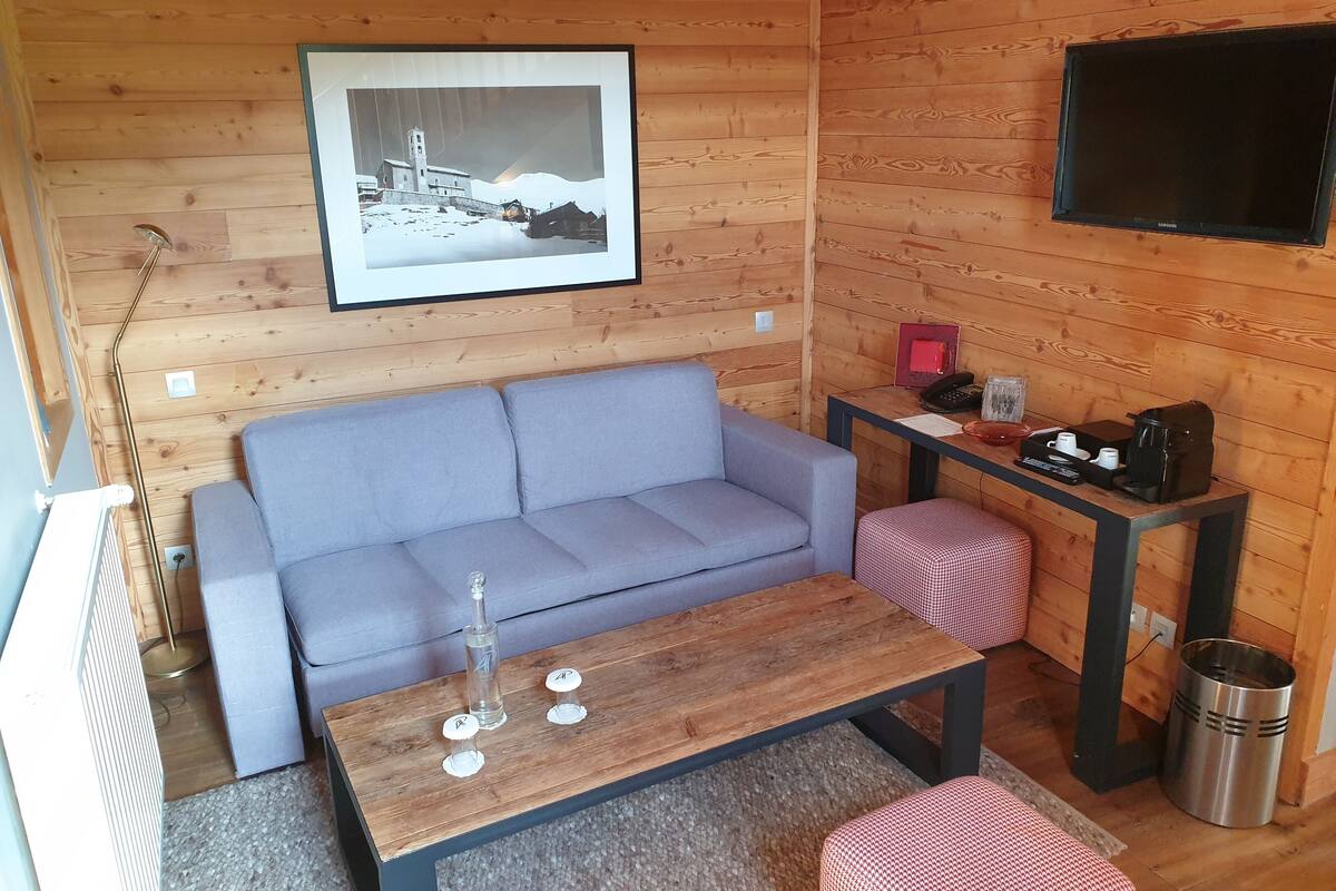 A cozy cabin interior with a grey sofa, a wooden coffee table, a small red and white checkered ottoman, a framed black and white photo on the wall, and a Samsung TV mounted above a wooden table.