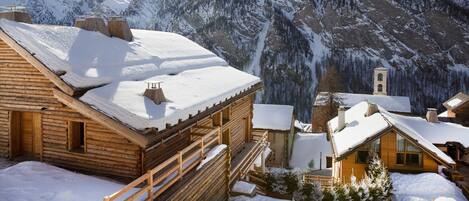 Una cabaña de madera con techo nevado, frente a montañas cubiertas de nieve y un cielo despejado.