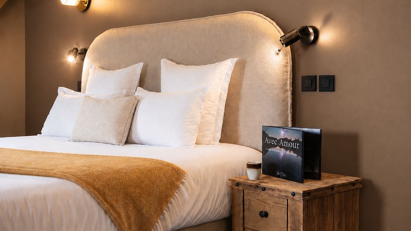 Una cama bien hecha con sábanas blancas y una manta marrón, una mesita de noche con un libro titulado 'Avec Amour', y una lámpara de pared.