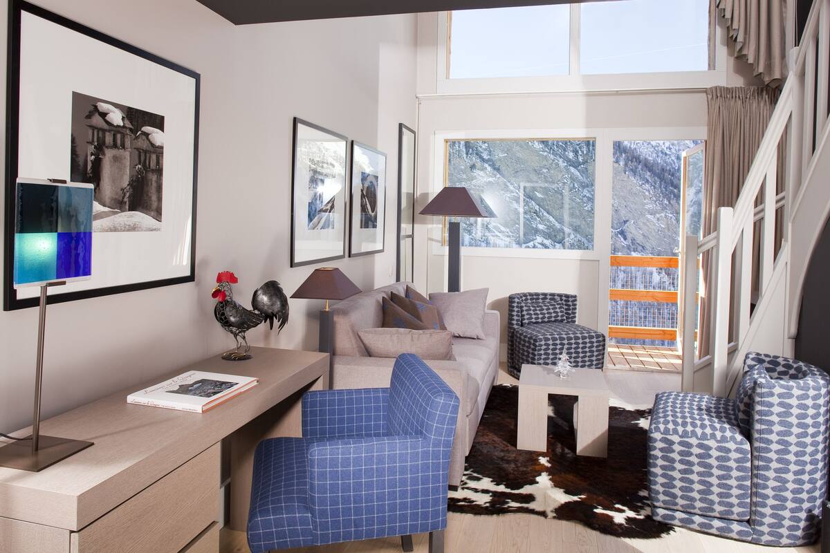 A modern living room with a sofa, armchairs, a coffee table, and a desk with a lamp. There are framed pictures on the wall and a view of snowy mountains through the windows.