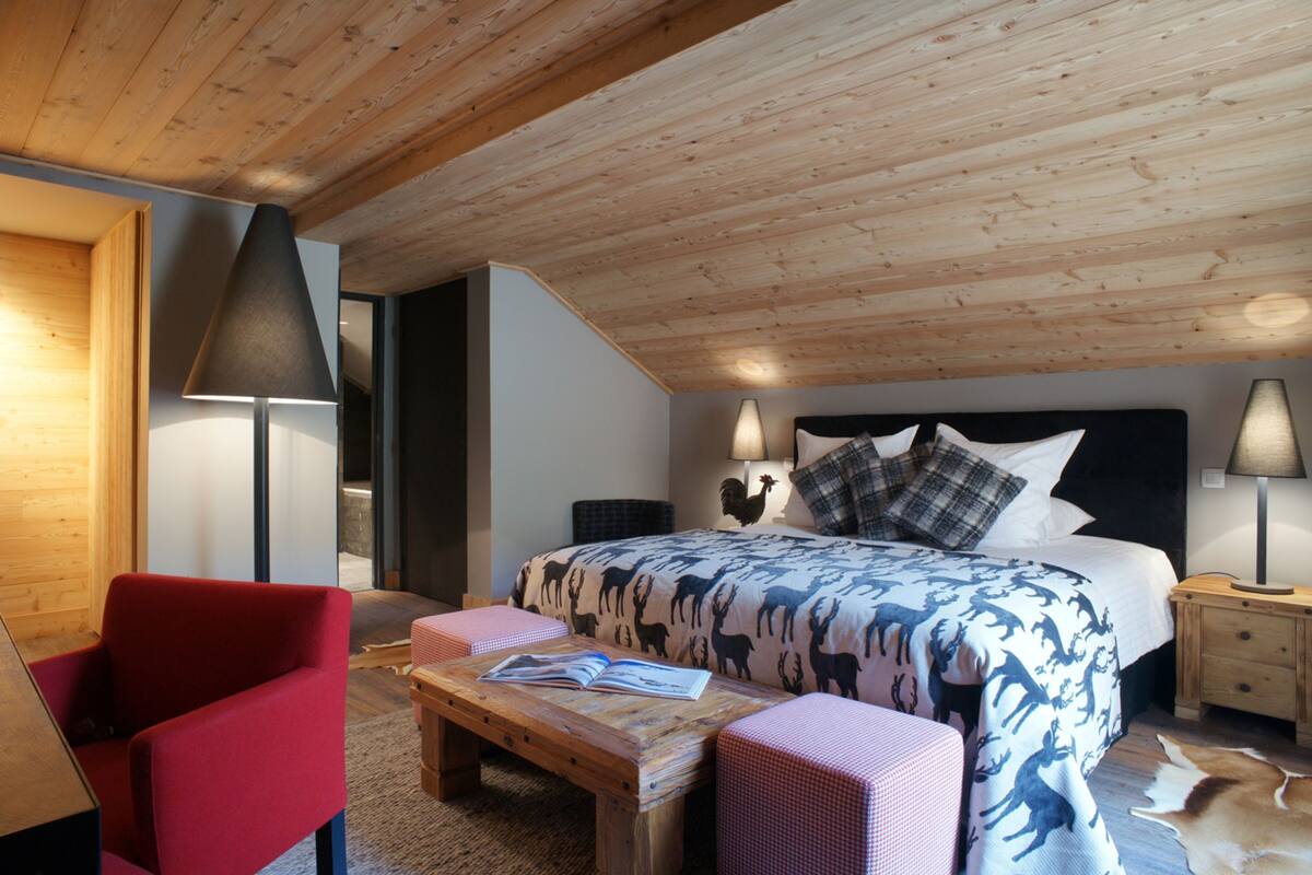 A bedroom with a wooden ceiling, a bed with patterned bedding, a red chair, a wooden table, and a nightstand with a lamp.