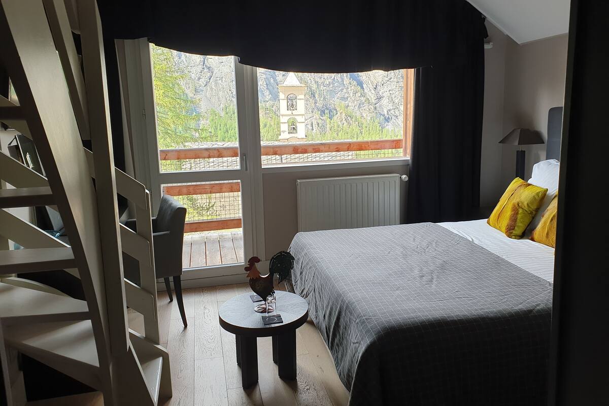 A bedroom with a bed, a small round table, a chair, and a view of a mountain and a tower through a large window.