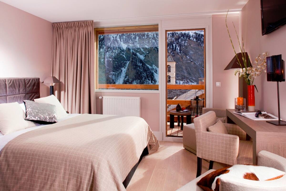 A hotel room with a large bed, a desk, a chair, and a view of a snowy mountain through a window.