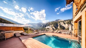 A wooden deck with a pool, lounge chairs, and a bar area overlooking a mountainous landscape. - Hotel & Spa L'Alta Peyra (Saint-Veran)