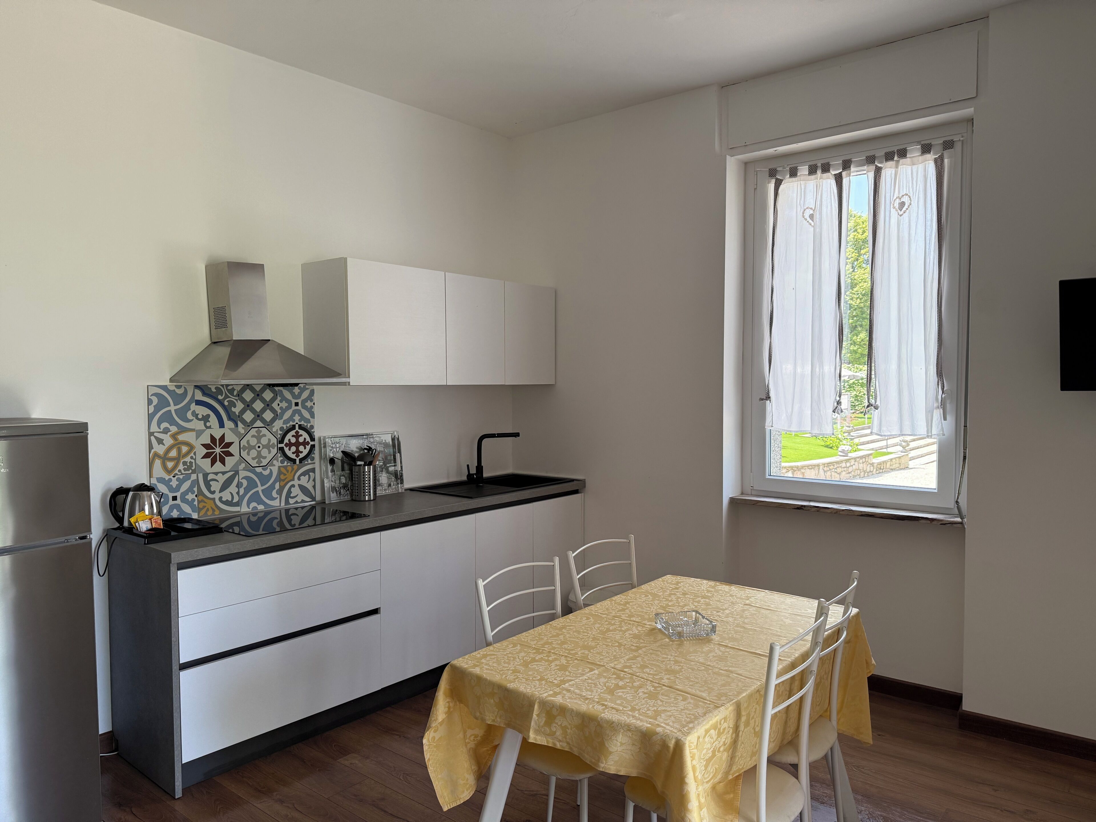 A compact kitchen with a refrigerator, stove, and white cabinets. A dining table with chairs is positioned near a window with curtains.