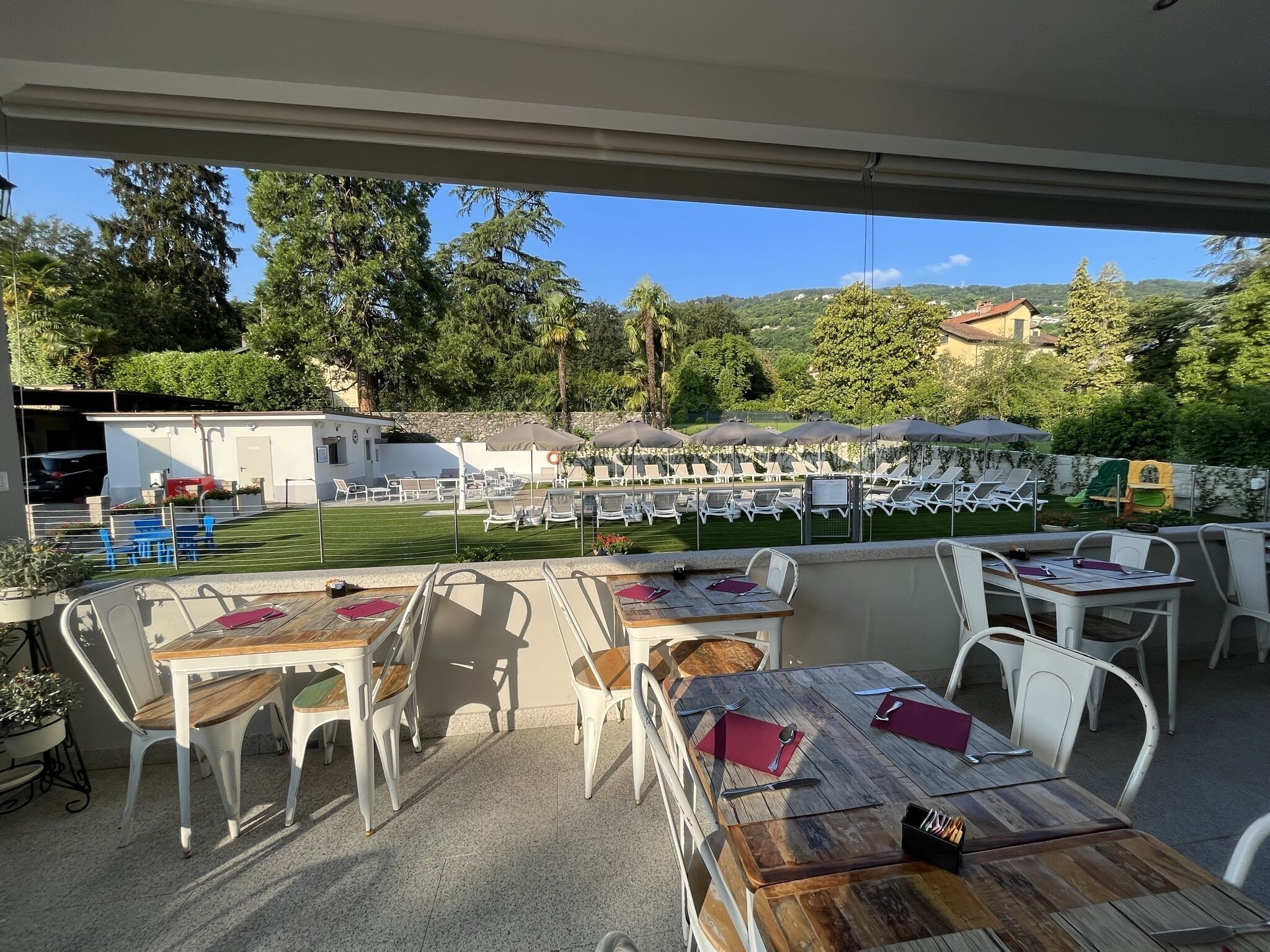 An outdoor patio with tables and chairs, a pool area with lounge chairs and umbrellas, and a view of the surrounding greenery and hills.