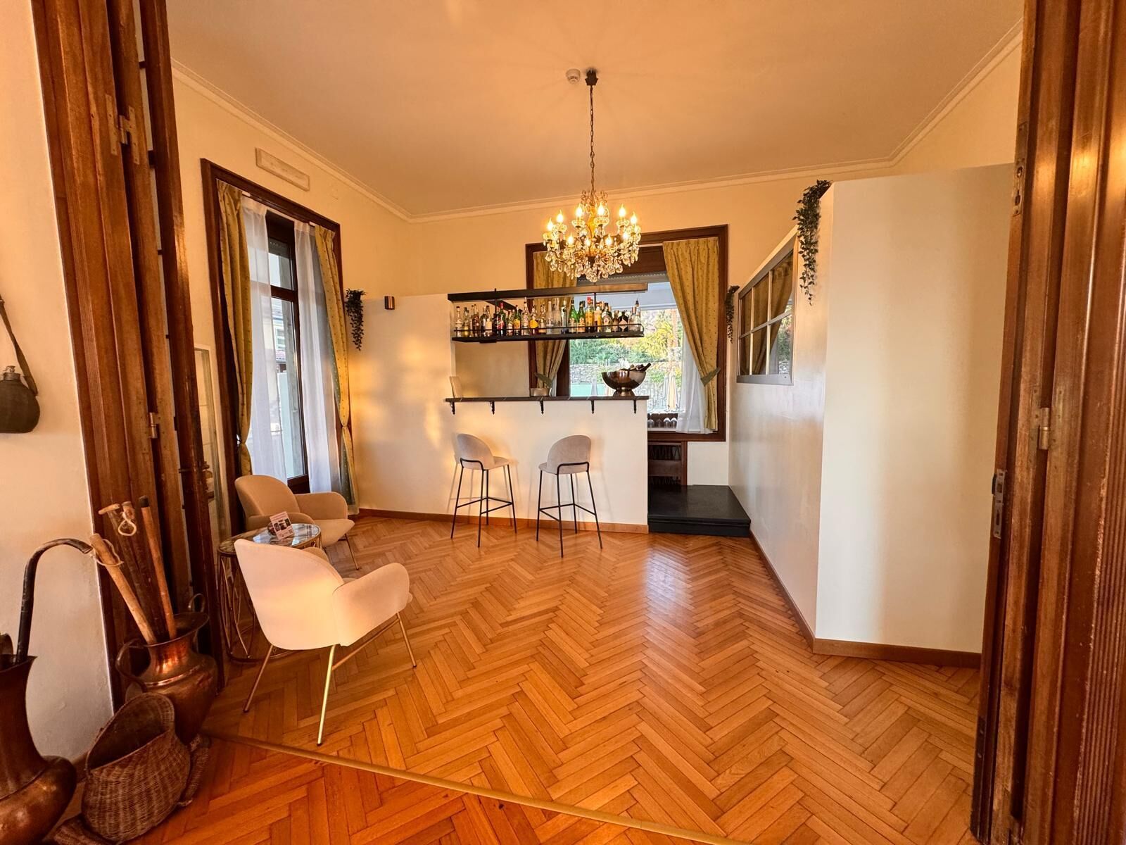 A room with a wooden herringbone floor, a chandelier, a bar area with bottles, and two white chairs.