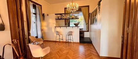 A room with a wooden herringbone floor, a chandelier, a bar area with bottles, and two white chairs.