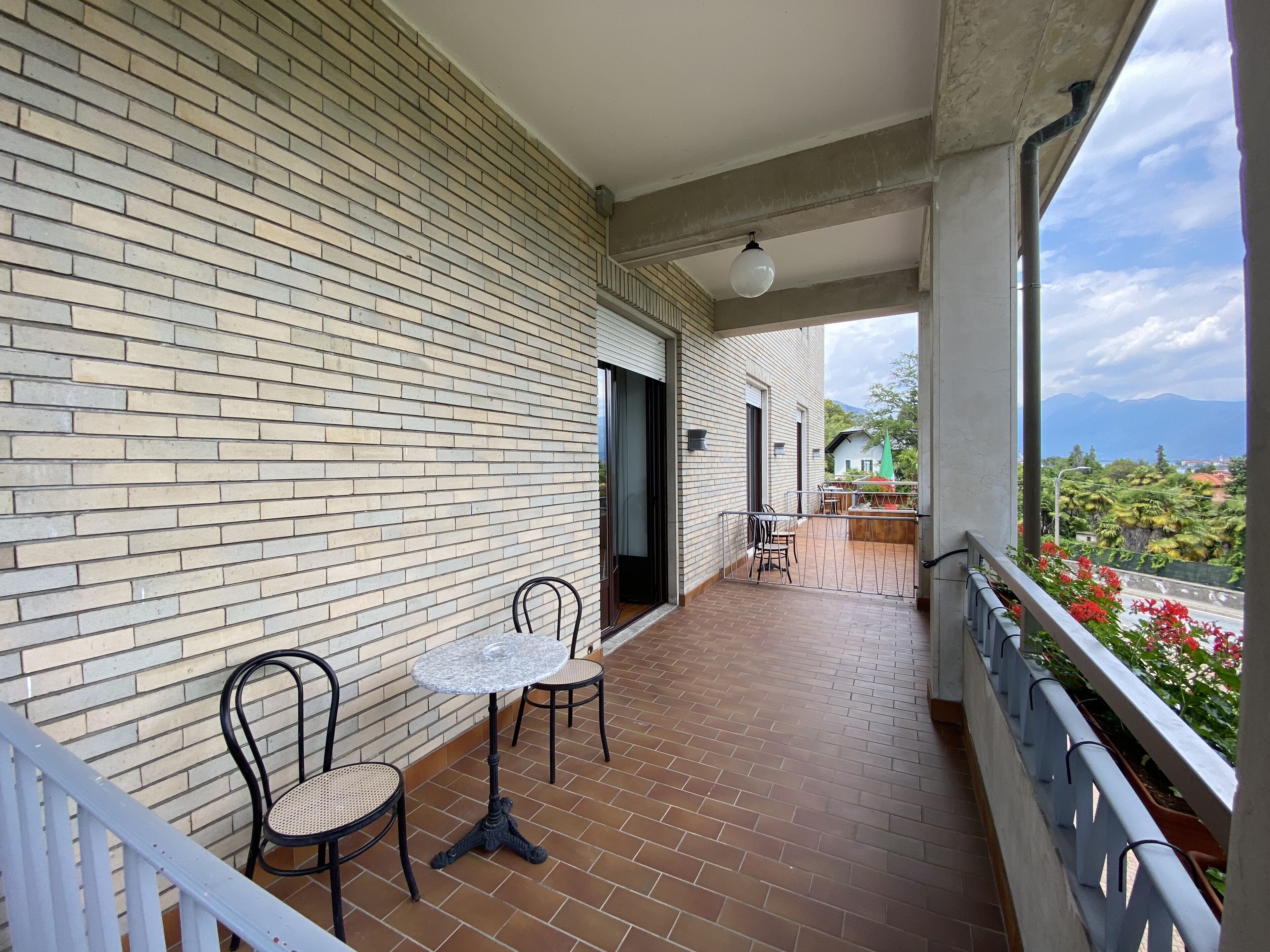 A balcony with a brick wall, two chairs, a small table, and a view of greenery and mountains.