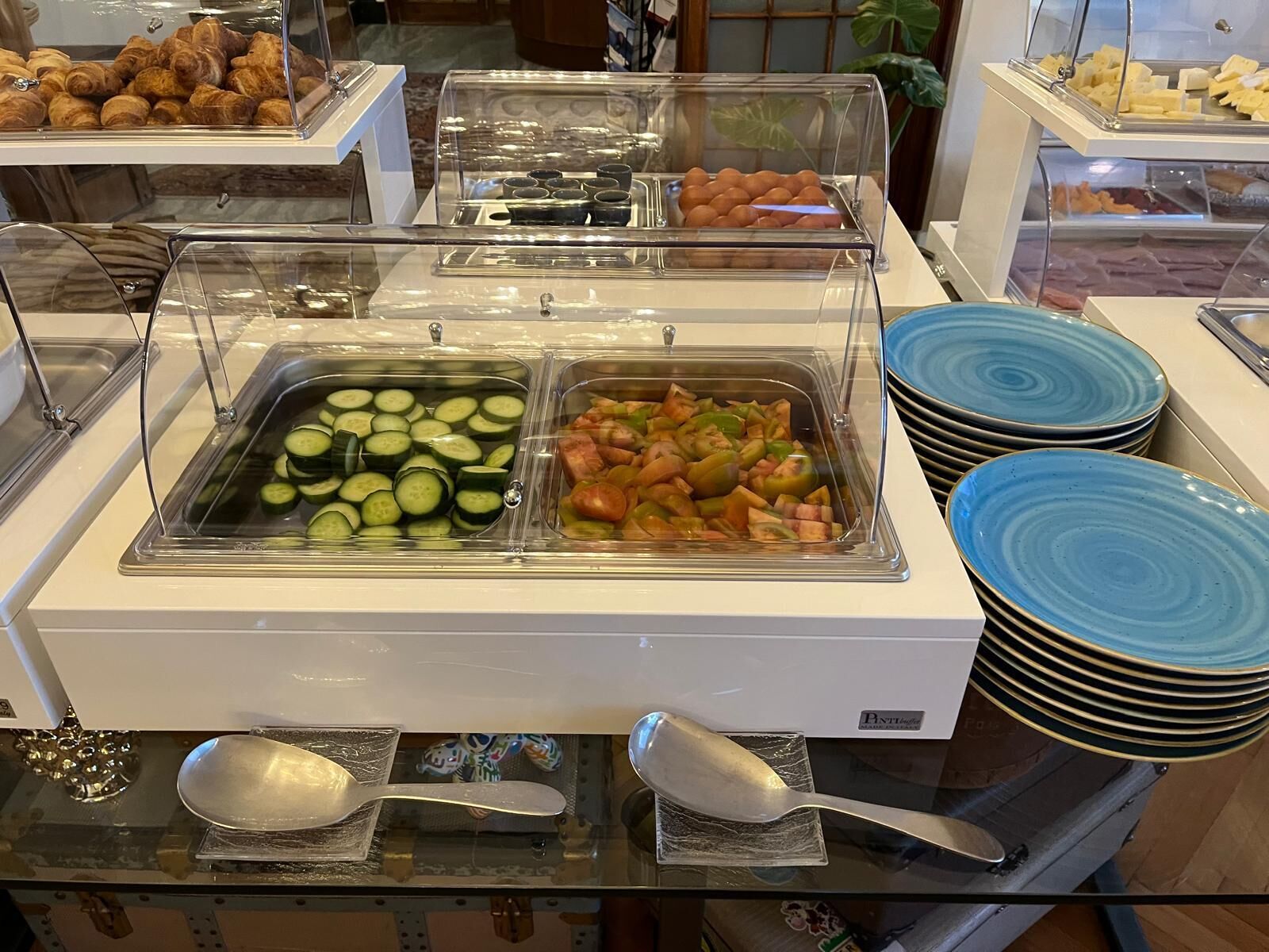 A buffet with clear plastic covers over food items, including sliced cucumbers, mixed vegetables, and pastries.