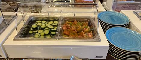 A buffet with clear plastic covers over food items, including sliced cucumbers, mixed vegetables, and pastries.
