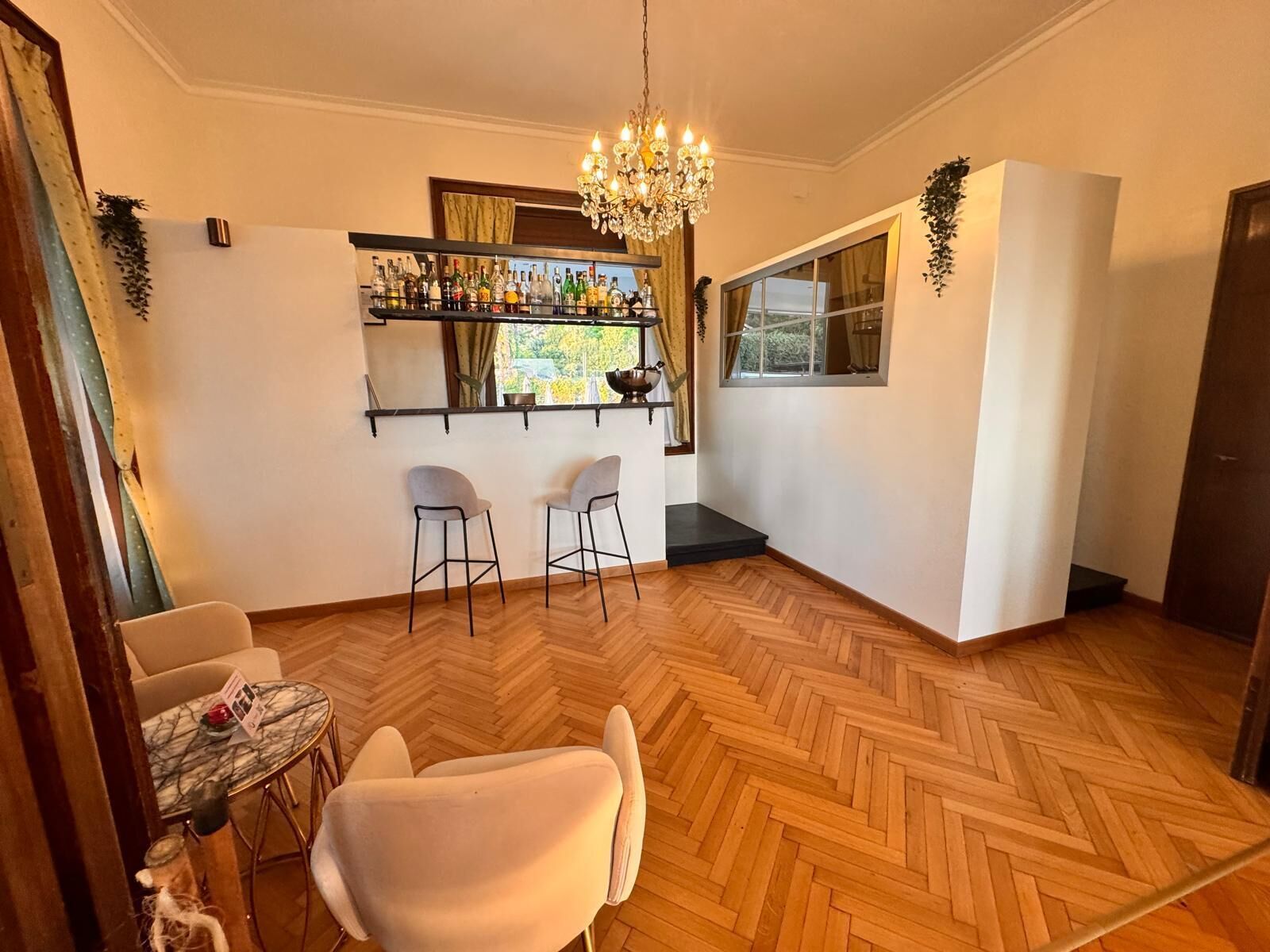 A room with a bar area, two bar stools, a chandelier, a dining table with chairs, and a herringbone wooden floor.