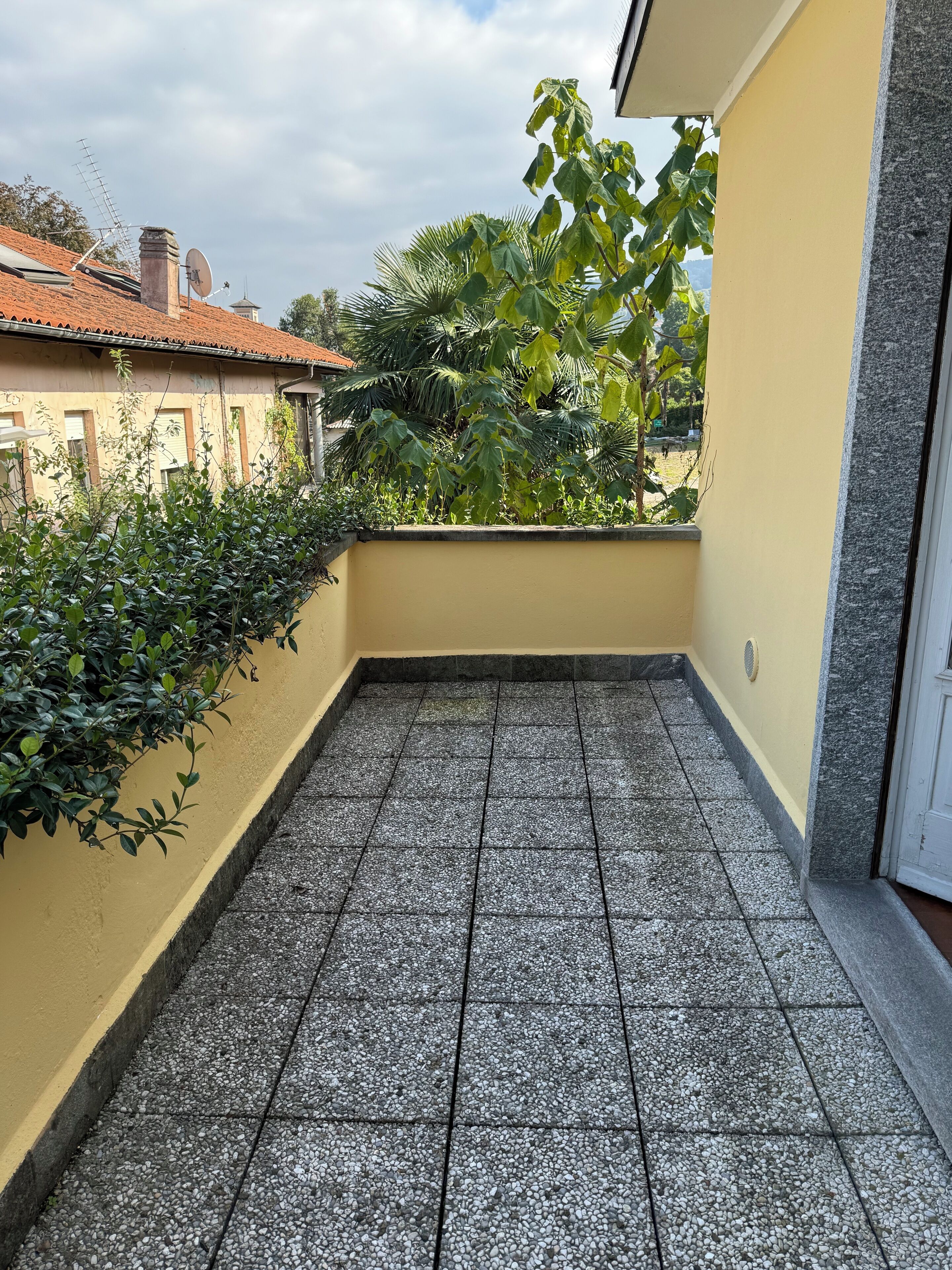 A balcony with tiled flooring, a yellow wall, and a view of a building with a red roof.