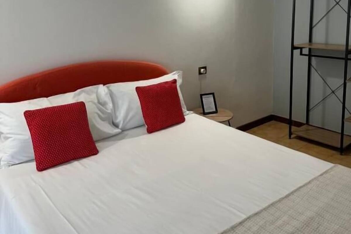 Una camera d'albergo con un letto, una testiera imbottita rossa e un corpo illuminante fissato al muro.