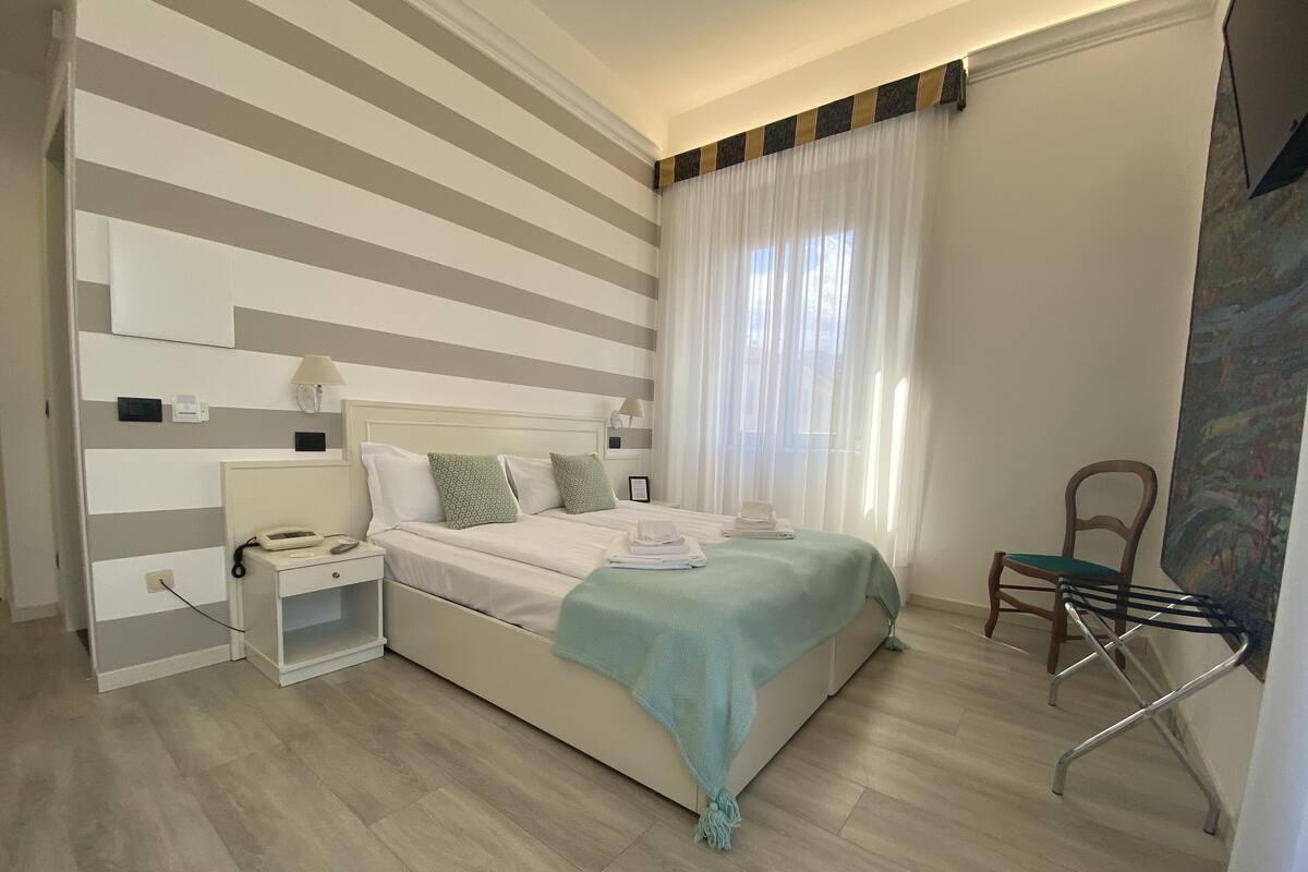 Camera d'albergo con parete a righe, un letto con coperta verde, un comodino con telefono e una sedia con portasciugamani.