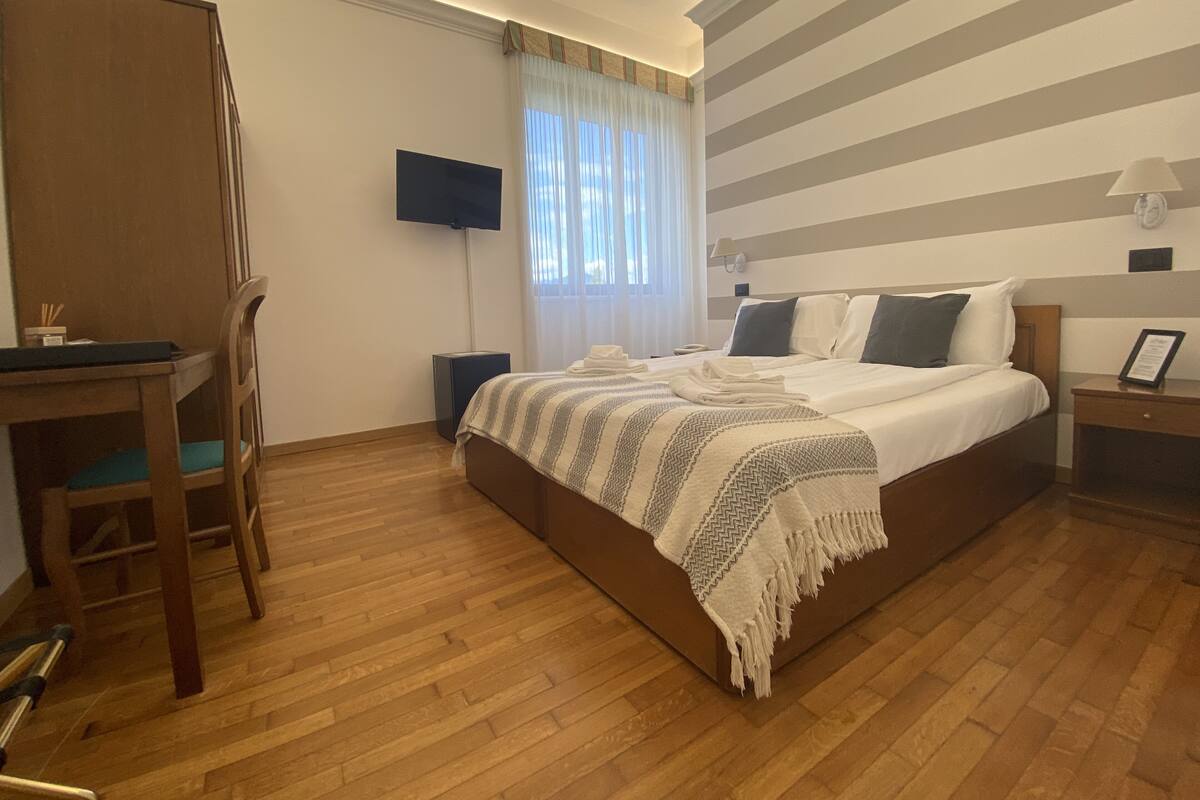 Una camera d'albergo con un letto grande, una scrivania con una sedia, una TV a schermo piatto, un comodino e una finestra con tende.