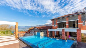 Una piscina infinita en la azotea con vistas a montañas, una terraza de madera y un edificio moderno con grandes ventanales.