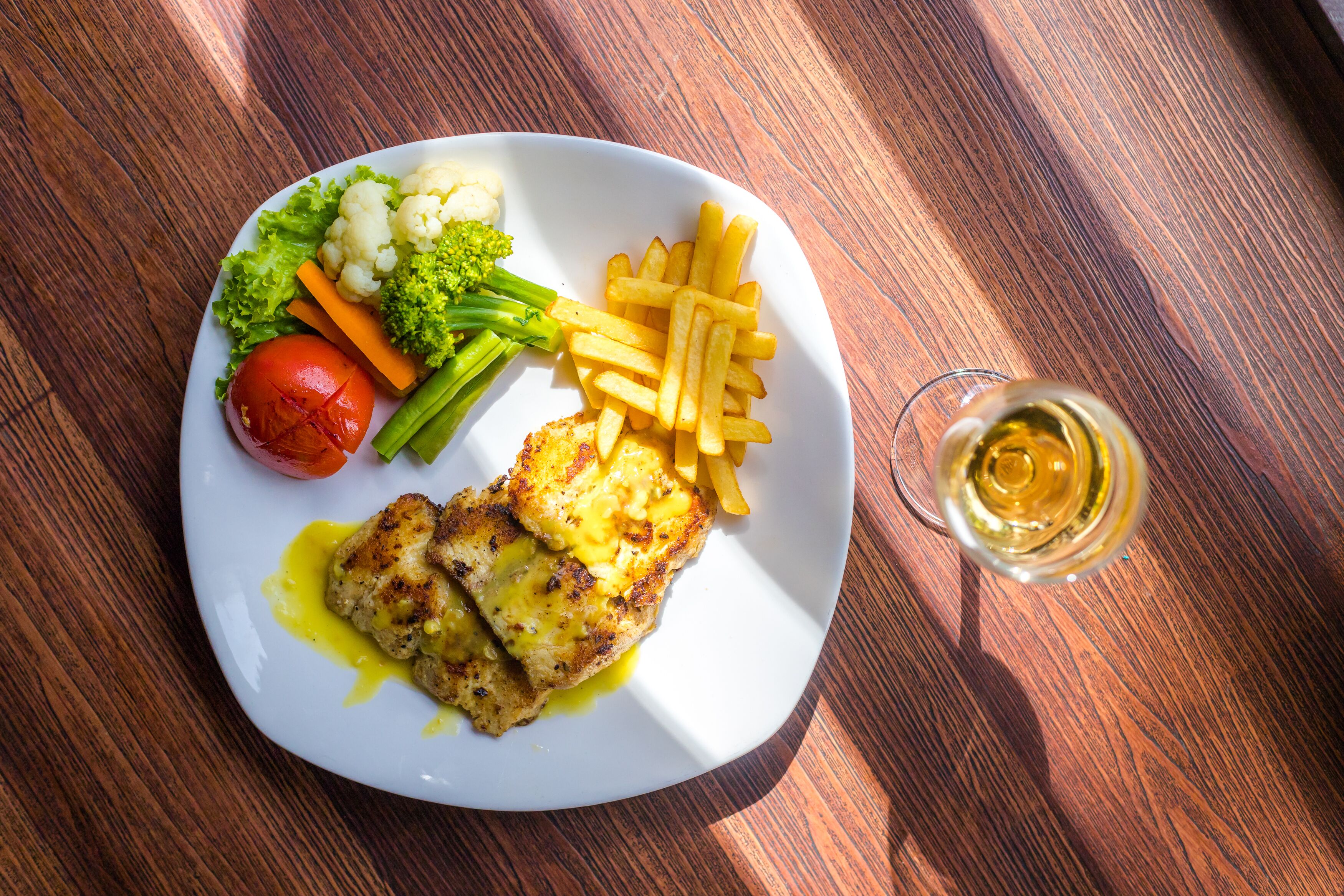 A plate with grilled chicken, French fries, and vegetables on a wooden table.