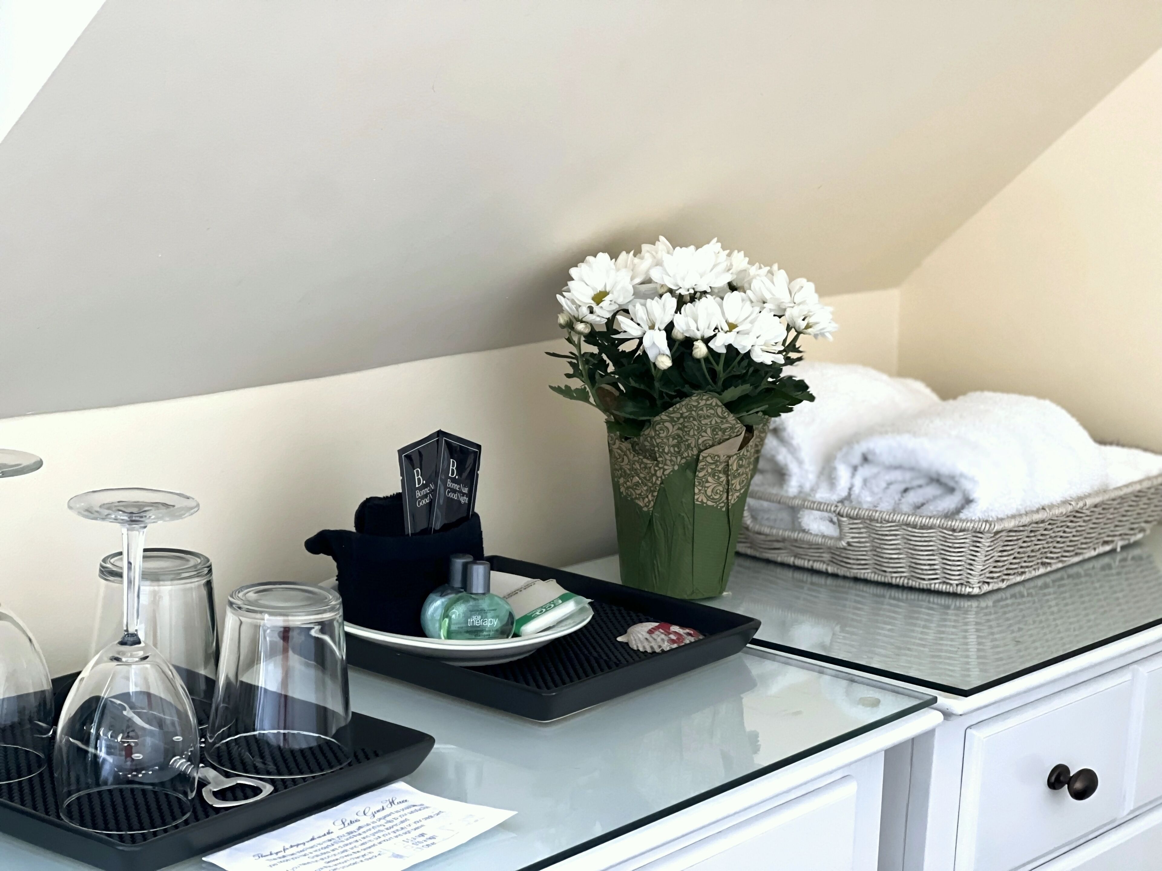 Habitación de hotel con una mesita de vidrio, una bandeja con copas de vino, un florero con flores blancas, una cesta con toallas y una bandeja con artículos de aseo.