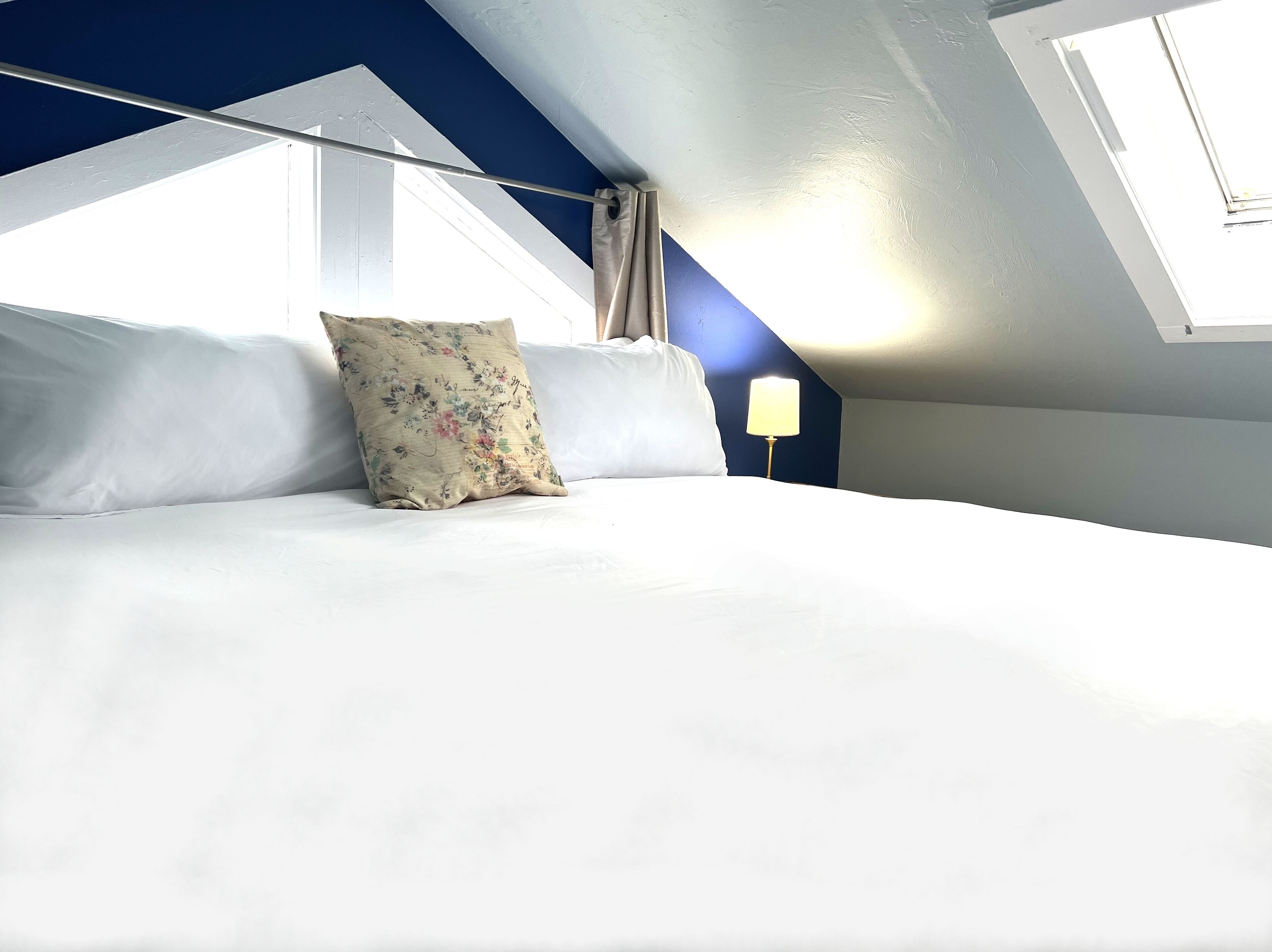 A bedroom with a sloped ceiling, a bed with white linens, a floral pillow, and a bedside lamp.