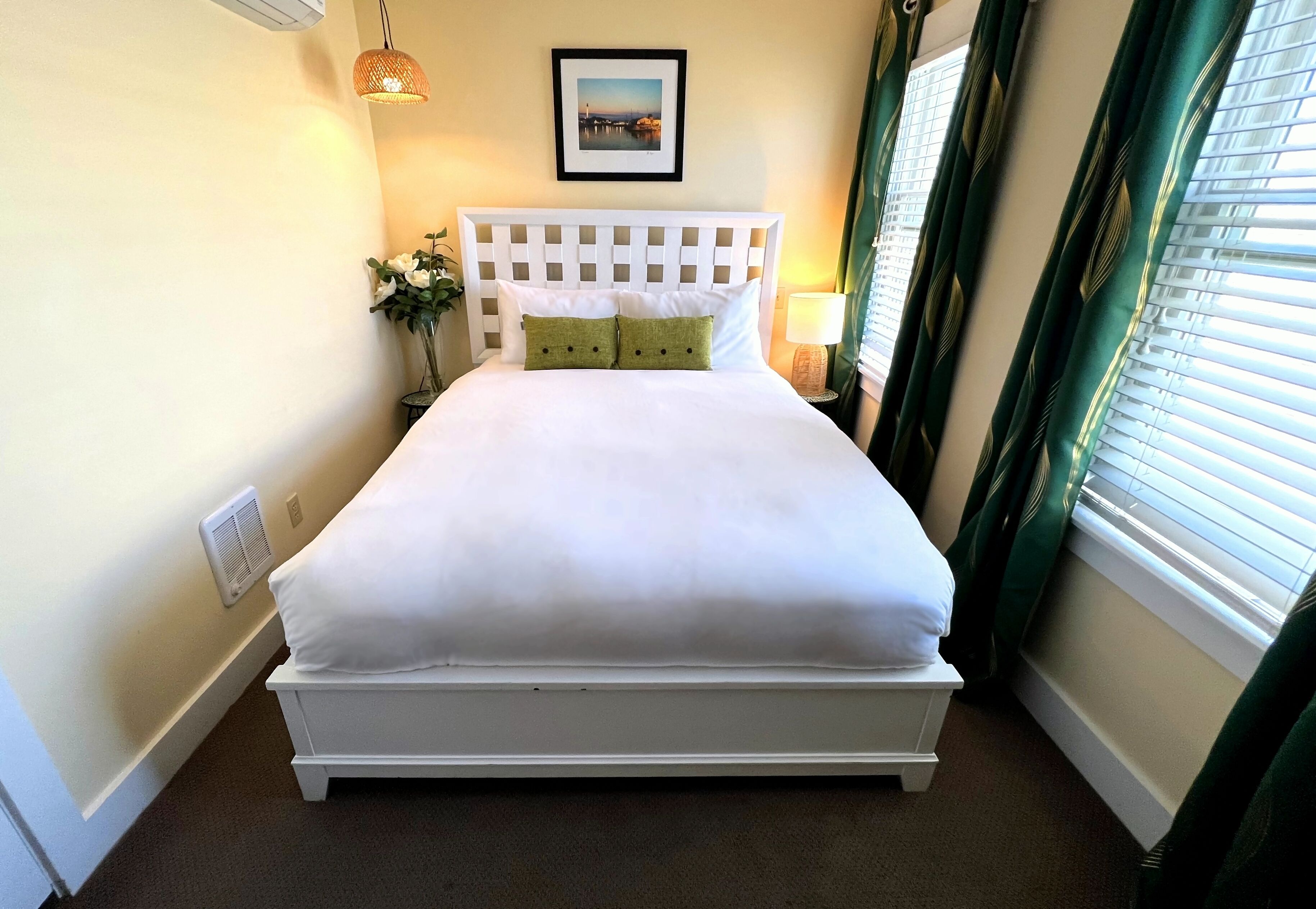 A neatly made bed with white linens and two pillows, a wall-mounted picture, a bedside lamp, and a window with curtains.